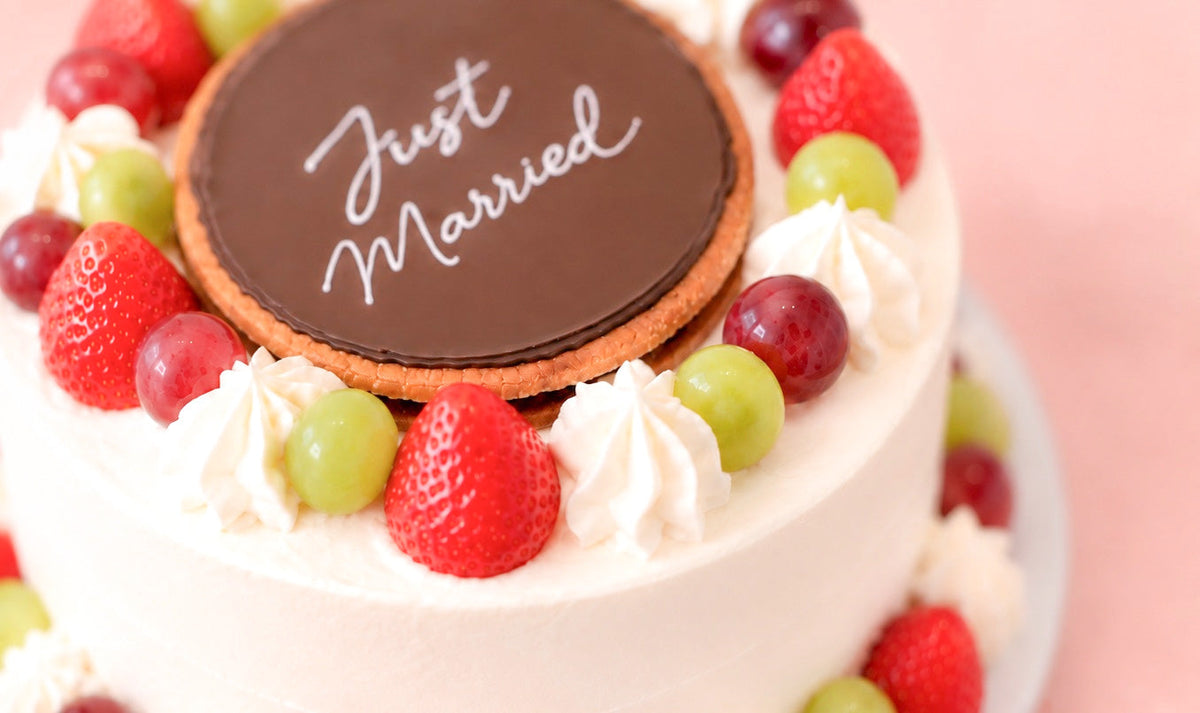 A heart-shaped pink frosted cake on a white plate, featuring a chocolate heart topper with the words in white script, set against a solid pink background.