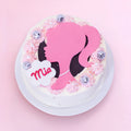 A round white-frosted cake on a white plate, decorated with pink piped rosettes, a large pink silhouette of a woman with black hair, a white cloud-shaped topper reading 'Mia', and several small silver disco-ball-style ornaments, set against a soft pink background.