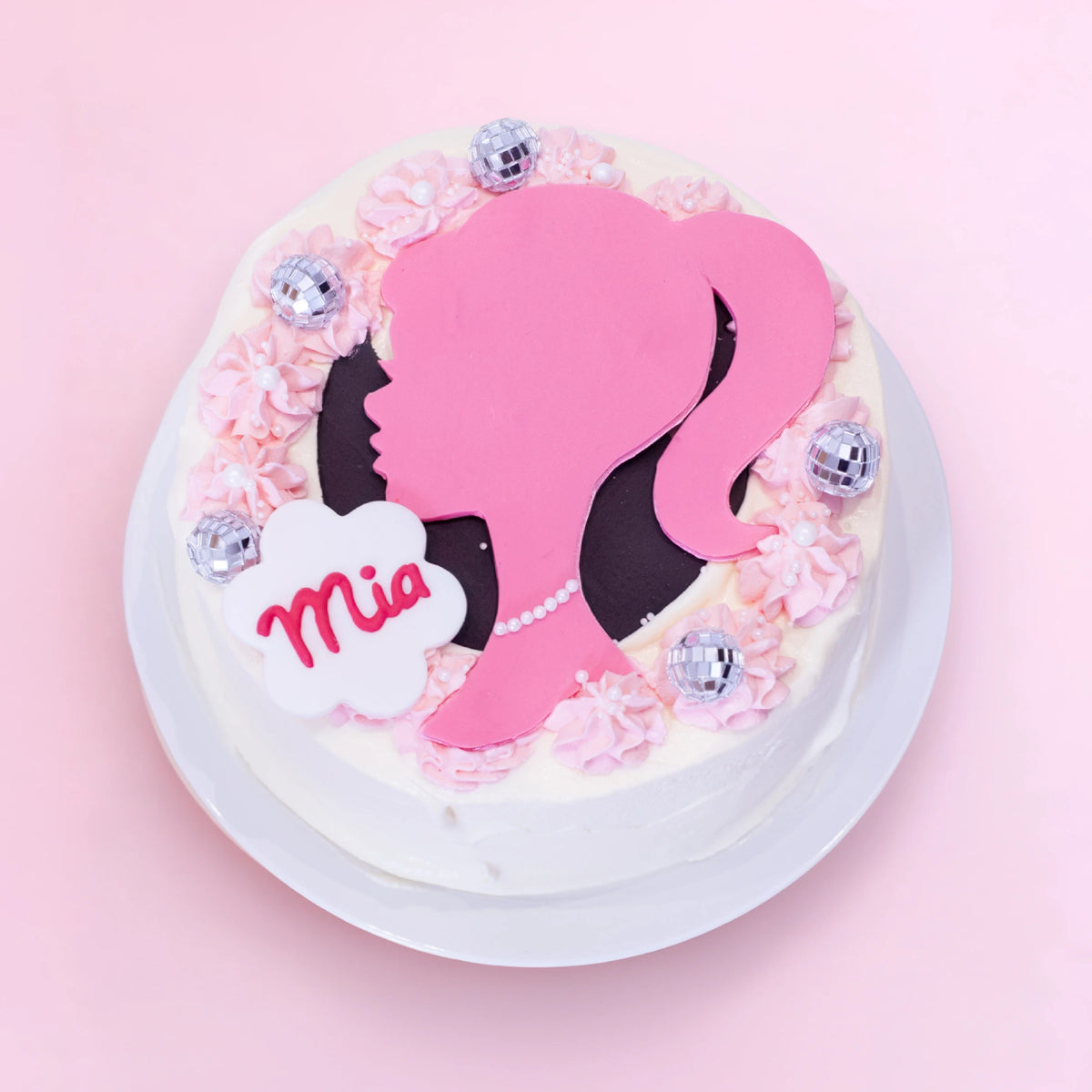 A round white-frosted cake on a white plate, decorated with pink piped rosettes, a large pink silhouette of a woman with black hair, a white cloud-shaped topper reading 'Mia', and several small silver disco-ball-style ornaments, set against a soft pink background.