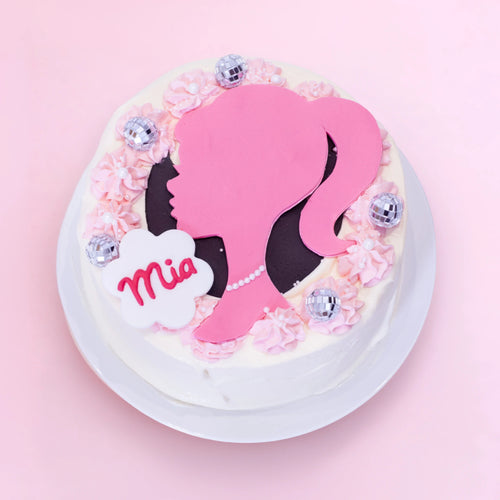 A round white-frosted cake on a white plate, decorated with pink piped rosettes, a large pink silhouette of a woman with black hair, a white cloud-shaped topper reading 'Mia', and several small silver disco-ball-style ornaments, set against a soft pink background.
