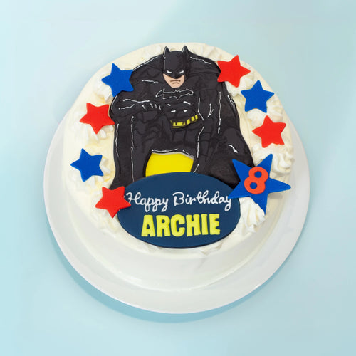 A round white-frosted birthday cake topped with a detailed Batman figure, red and blue star decorations, and a blue plaque reading 'Happy Birthday ARCHIE 8' on a light blue background.