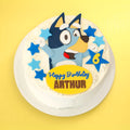 A round white frosted birthday cake topped with a fondant Bluey character, blue stars, and a yellow sign reading 'Happy Birthday ARTHUR', shown on a yellow background.