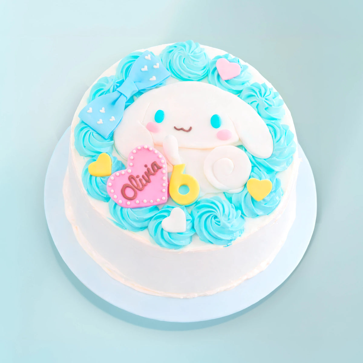 A round white cake decorated with light blue rosette frosting, a large white bunny face, a blue bow, a pink heart-shaped plaque reading 'Olivia', and yellow heart accents, shown on a light blue background.