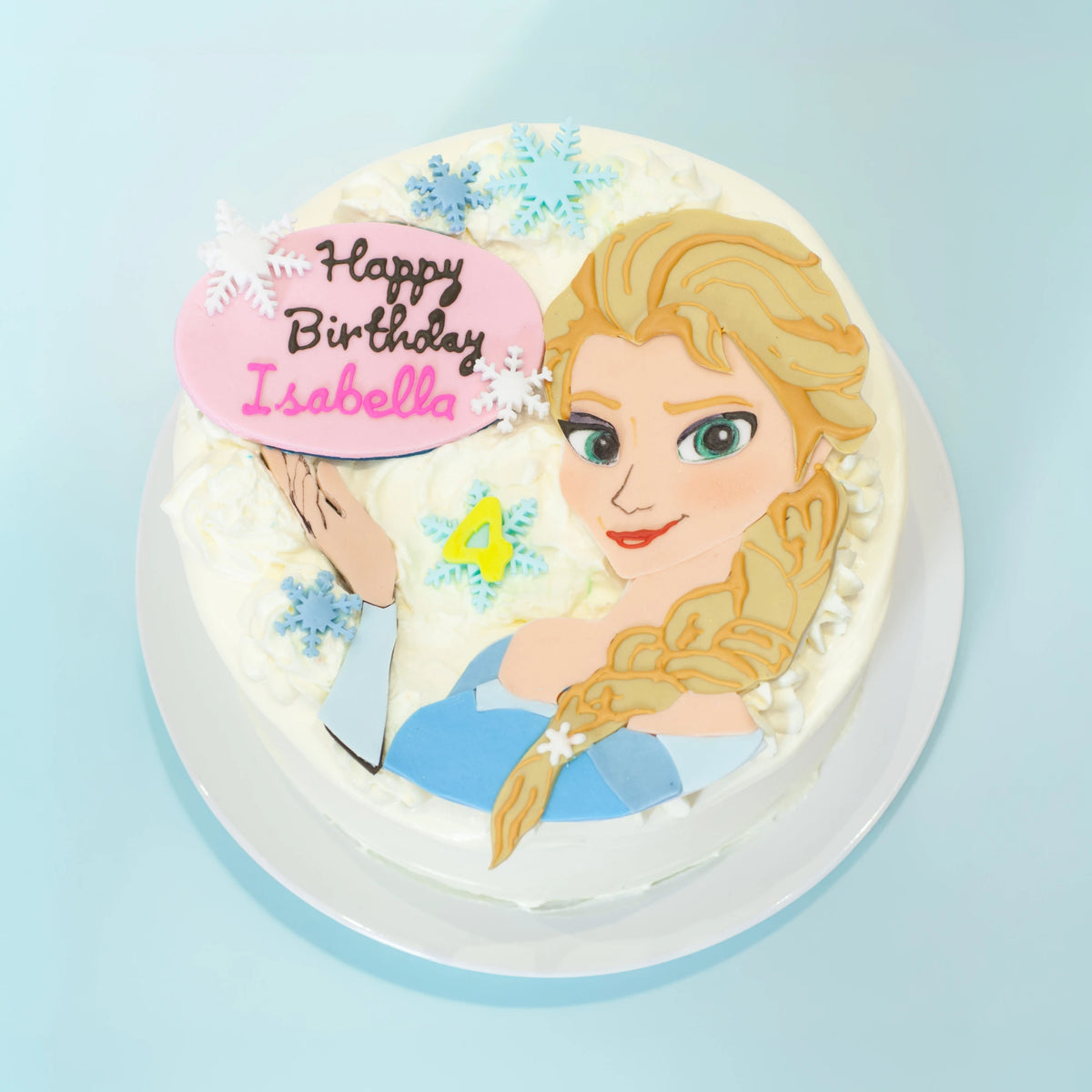 A round white-frosted birthday cake featuring a printed illustration of Elsa from Frozen, blue snowflake decorations, and a pink heart-shaped topper that reads 'Happy Birthday Isabella' with a yellow number 4.