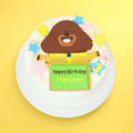 A round birthday cake decorated with a brown dog Hey Duggee character on top, colourful star sprinkles, and a green sign that reads 'Happy Birthday Harper', shown on a white plate against a bright yellow background.