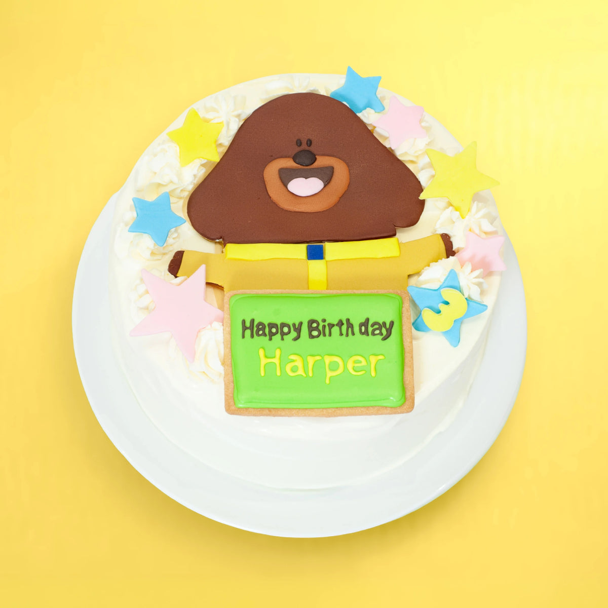 A round birthday cake decorated with a brown dog Hey Duggee character on top, colourful star sprinkles, and a green sign that reads 'Happy Birthday Harper', shown on a white plate against a bright yellow background.