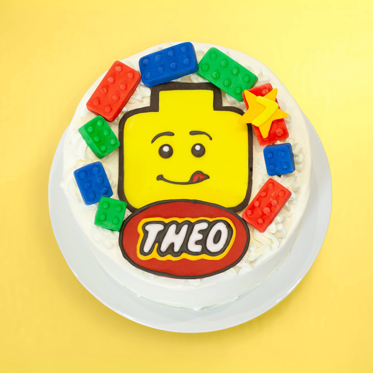 A round white frosted cake decorated like a LEGO-themed birthday cake, featuring a large yellow LEGO minifigure head with a smiling face, colorful red, green, and blue LEGO bricks, and a red oval with the name 'THEO' in white. The cake sits on a white plate against a solid yellow background.