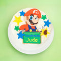 A round birthday cake decorated with a Mario character design, colorful icing stars, and a green sign that reads 'Happy Birthday Jude,' shown on a white plate against a light green background.