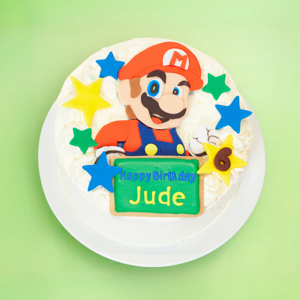 A round birthday cake decorated with a Mario character design, colorful icing stars, and a green sign that reads 'Happy Birthday Jude,' shown on a white plate against a light green background.