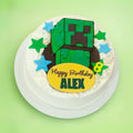 A round white-frosted birthday cake topped with a green Minecraft-style block character, blue and green star decorations, and a yellow oval plaque reading 'Happy Birthday ALEX', shown on a light green background.