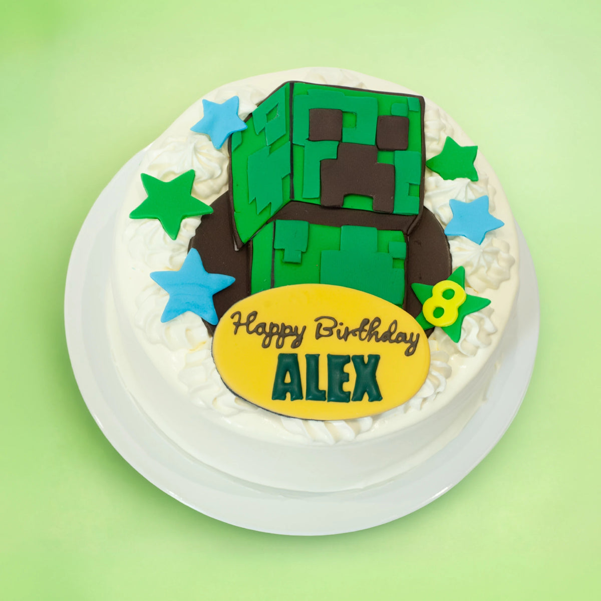A round white-frosted birthday cake topped with a green Minecraft-style block character, blue and green star decorations, and a yellow oval plaque reading 'Happy Birthday ALEX', shown on a light green background.
