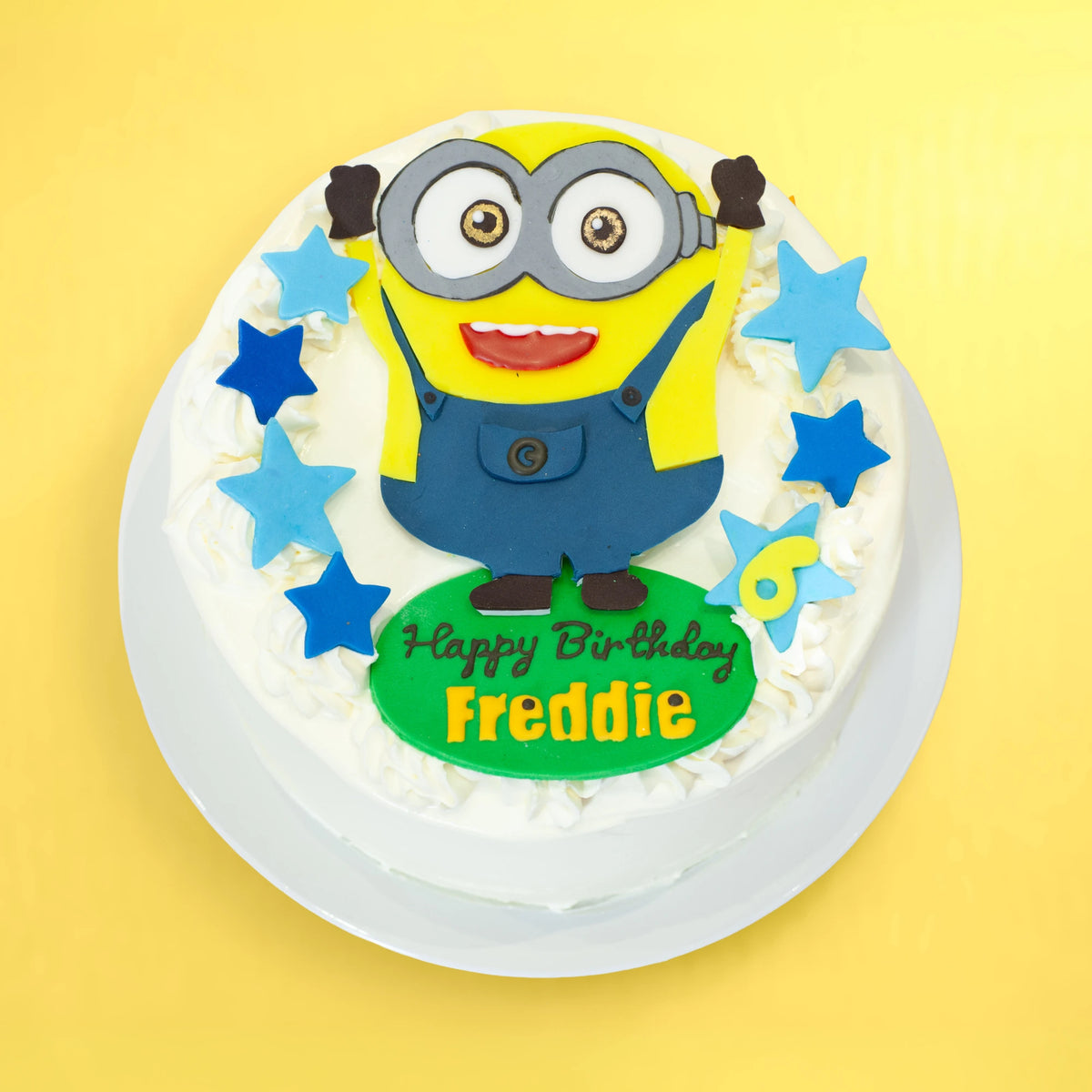A round white-frosted birthday cake topped with a Minions character decoration, blue and light blue star shapes, and a green plaque reading 'Happy Birthday Freddie', shown on a yellow background.