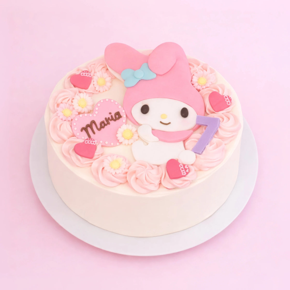 A round white cake decorated with pink frosting rosettes, small daisy flowers, pink hearts, and a large pink character topper resembling My Melody, with the name 'Maria' on a pink heart and a purple number 7, set on a white plate against a pink background.