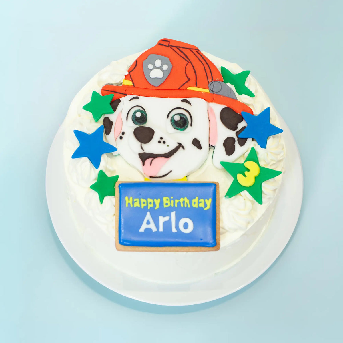 A round white frosted birthday cake decorated with a Dalmatian dog face wearing an orange firefighter helmet, colorful star accents, and a blue sign that reads 'Happy Birthday Arlo', shown on a light blue background.