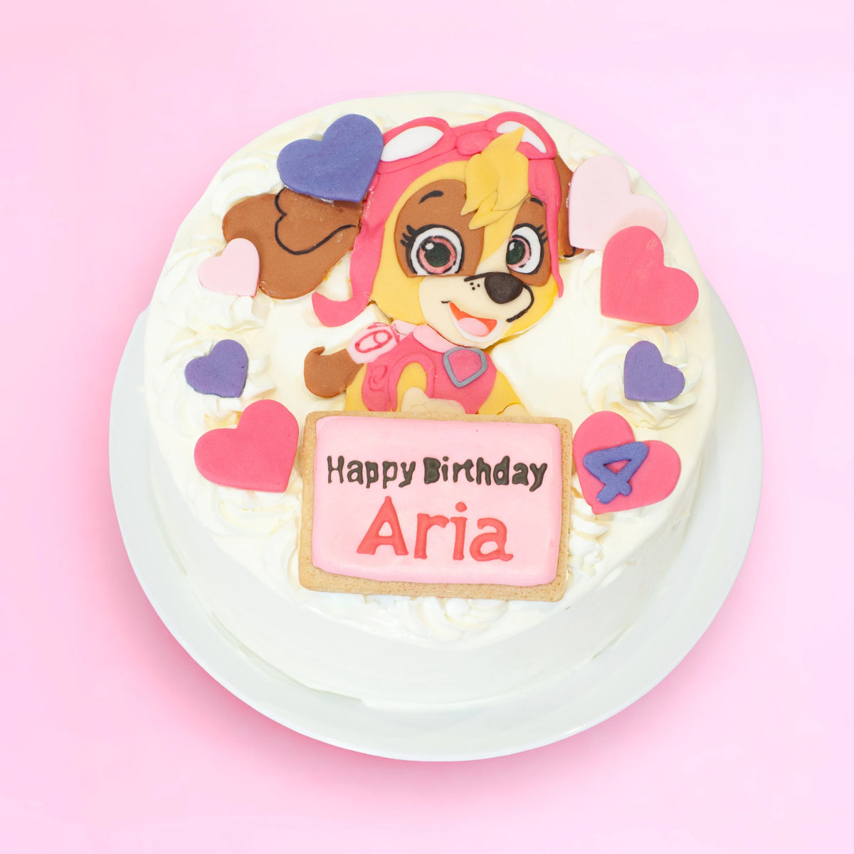 A round white-frosted birthday cake on a white plate, decorated with a Skye character fondant topper, pink and purple heart accents, and a pink sign reading 'Happy Birthday Aria' on a light pink background.
