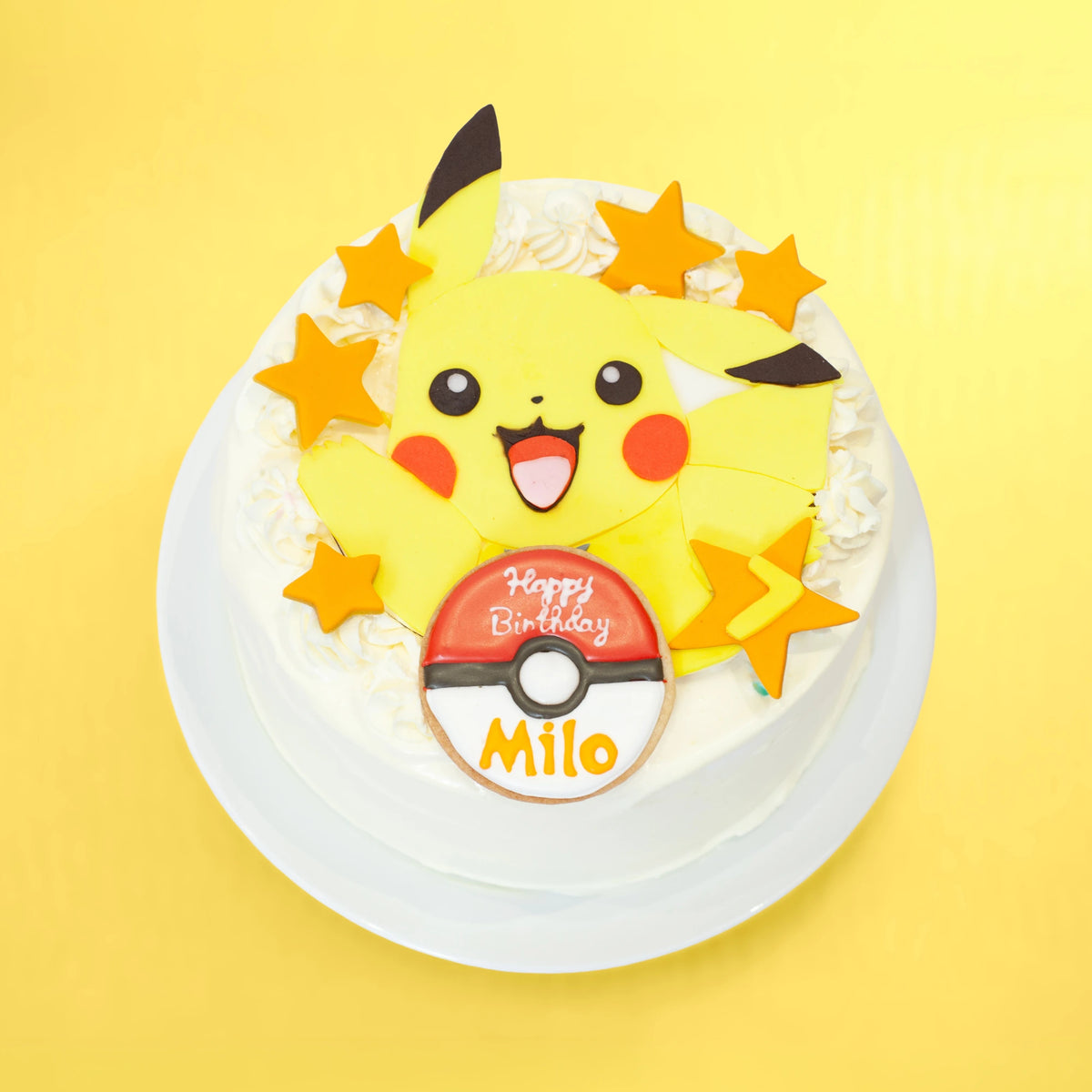 A round white frosted birthday cake topped with a large yellow Pikachu figure, orange star decorations, and a red-and-white Pokéball topper reading 'Happy Birthday Milo,' set on a yellow background.