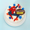 A round white-frosted birthday cake topped with a Spider-Man figure, blue and orange star decorations, and a brown oval sign that reads 'Happy Birthday OSCAR', shown on a light blue background.