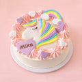 A round white cake decorated with a pastel unicorn face, rainbow mane, pink and white piped rosettes, heart-shaped toppers, and a purple heart that reads 'Mika' on a pink background.